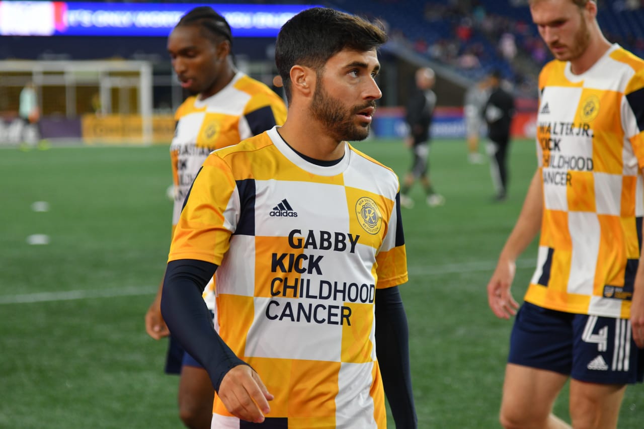 Revolution midfielder Carles Gil wearing a #KickChildhoodCancer training top during pregame warmups, with the top featuring the name of a local pediatric cancer patient from Boston Children's Hospital.